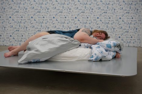 A person lying on a bed on a metal platform. The blankets are white with blue patterns, as is the wallpaper.