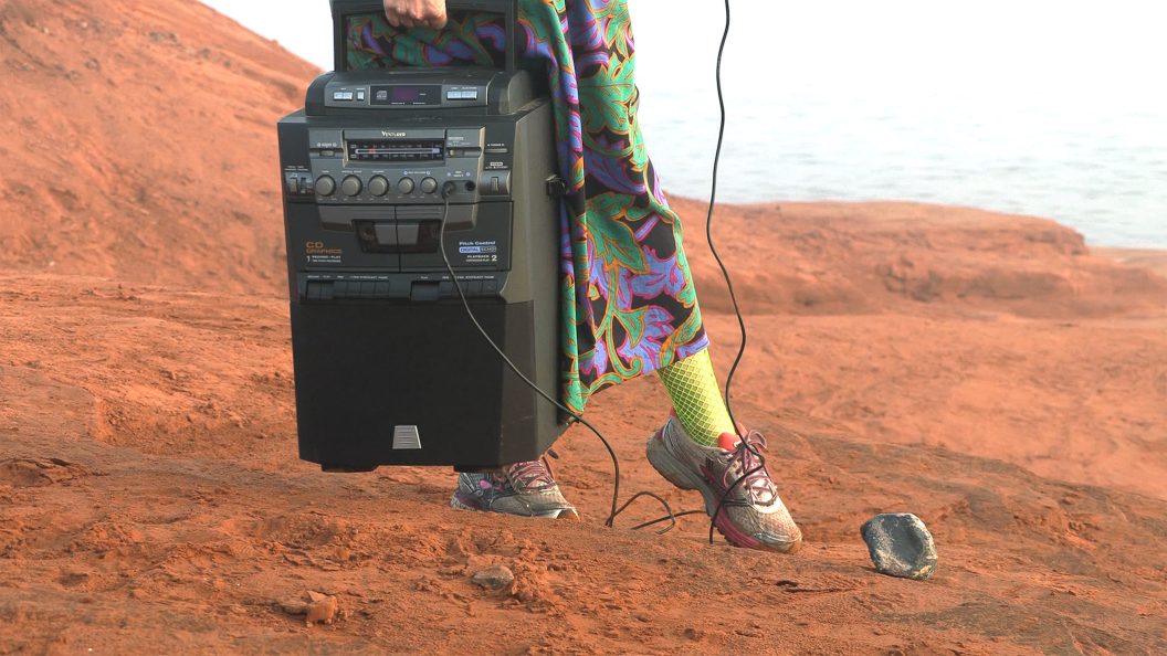 A person dressed in brightly colored patterned fabrics stands on a red sandbank, a portable stereo in hand.